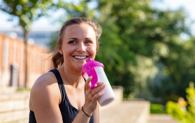 Vrouw in sportkleding heeft berekend hoeveel gram eiwitten ze per dag nodig heeft en drinkt een eiwitshake na haar training.