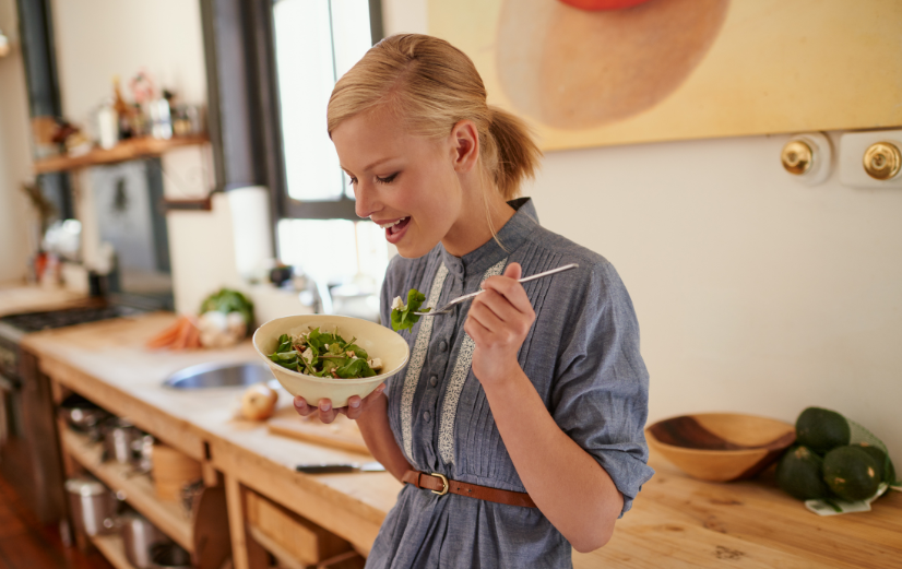 Vrouw staat in de keuken en eet een salade met groene bladgroenten, een bron van tocoferolen, vitamine E en antioxidanten.